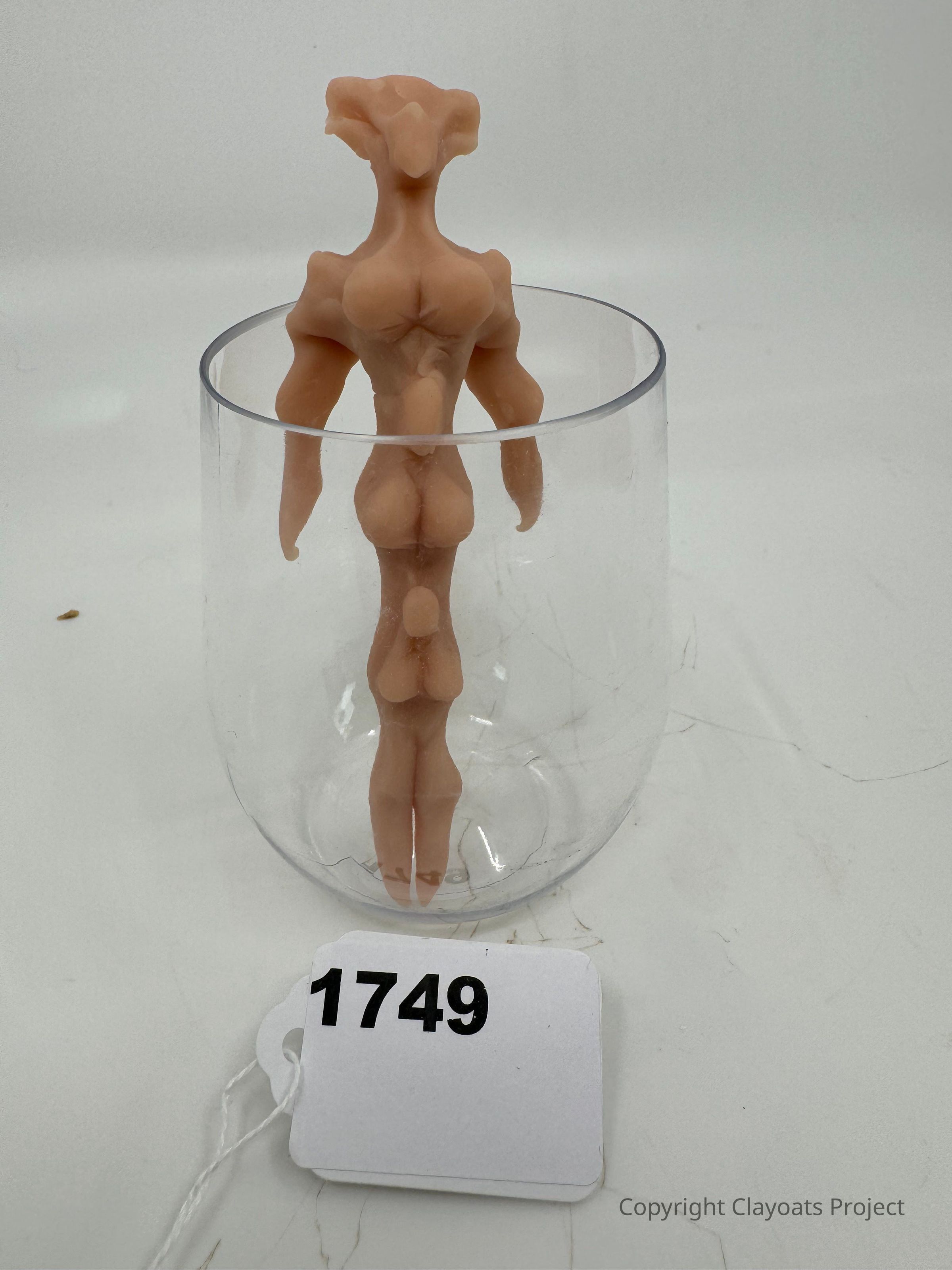 The image shows a small, abstract humanoid figure made of a pinkish material standing upright inside a transparent glass, positioned on a white surface. Its form seems muscular and stylized, lacking distinct facial features, with exaggerated proportions emphasizing the shoulders and torso, blending an organic and geometric aesthetic. The glass serves as a platform or enclosure, highlighting the figure's design and craftsmanship.