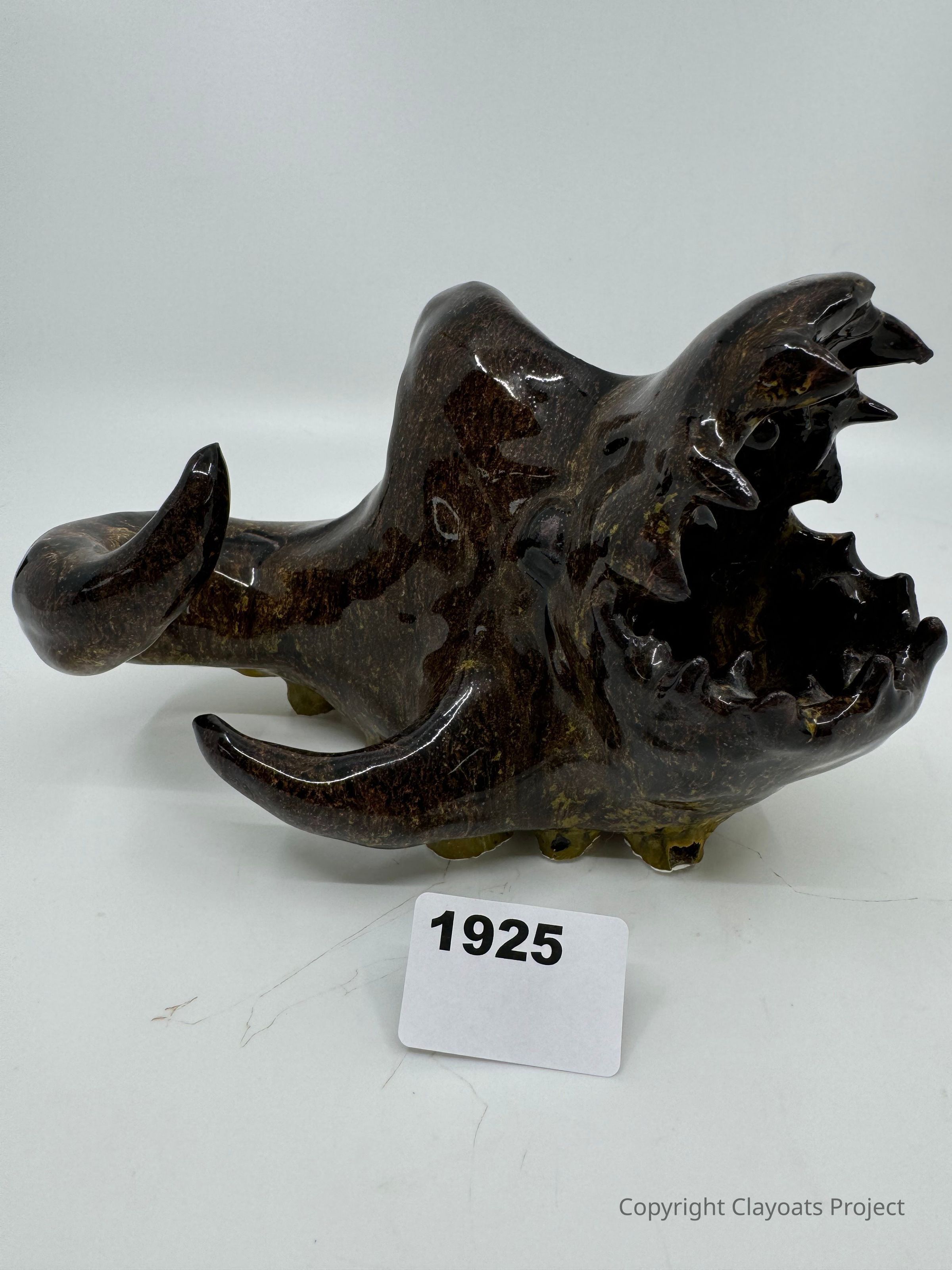 The image shows a glossy, dark sculpture with a rough, animal-like appearance, featuring prominent sharp teeth and a curved tail. The sculpture is placed against a plain white background, The texture of the sculpture resembles polished stone or ceramic, and it has an abstract, possibly mythical design. The lighting highlights its shiny surface, casting subtle reflections and shadows. Its unique and artistic form suggests it might be a decorative or