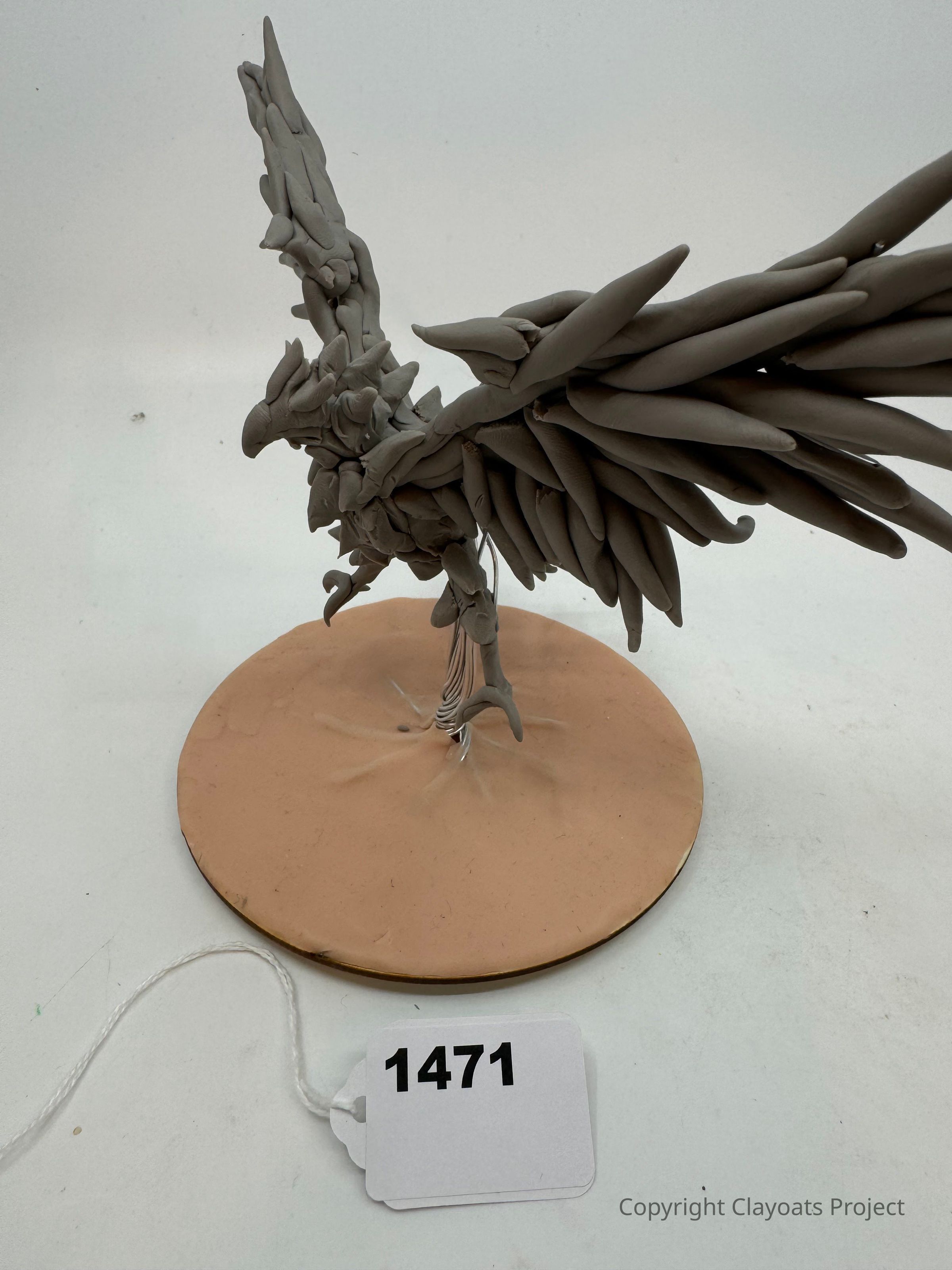 The image depicts a clay sculpture of a bird in flight, mounted on a circular base. The bird's intricate design features long, pointed feathers extending dramatically from its wings and body, giving it a dynamic, expressive appearance. The sculpture, crafted with a neutral tone clay, emphasizes the textures and forms of the bird's anatomy, capturing a sense of movement. The base, matching the clay's color, provides stability and contrast, highlighting the sculpture's intricate details