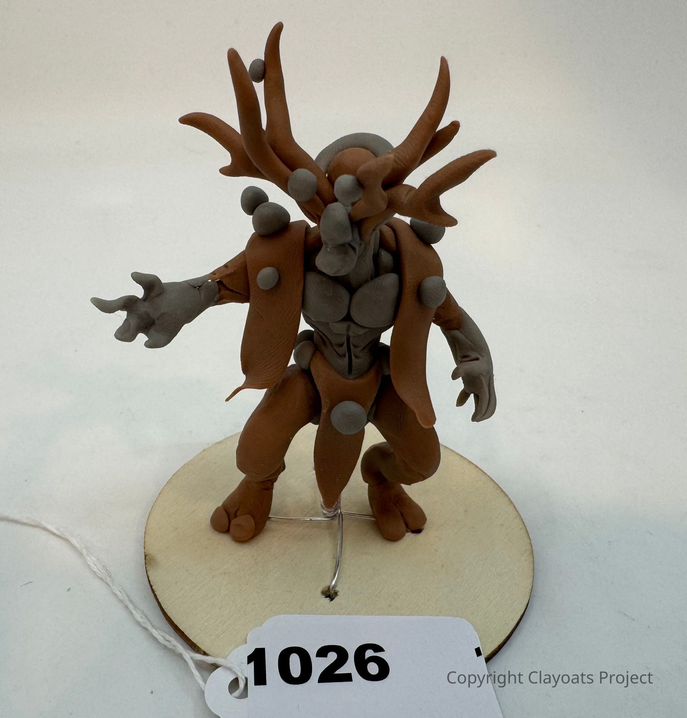 The image shows a sculpted clay figure resembling a fantastical creature, featuring a humanoid form with exaggerated limbs and antler-like appendages. The creature is crafted in two shades, brown and gray, with spherical accents adorning its body. It stands on a round wooden base, The background is plain and neutral, highlighting the intricate and imaginative details of the sculpture's design and posture, which suggest movement or an impending action