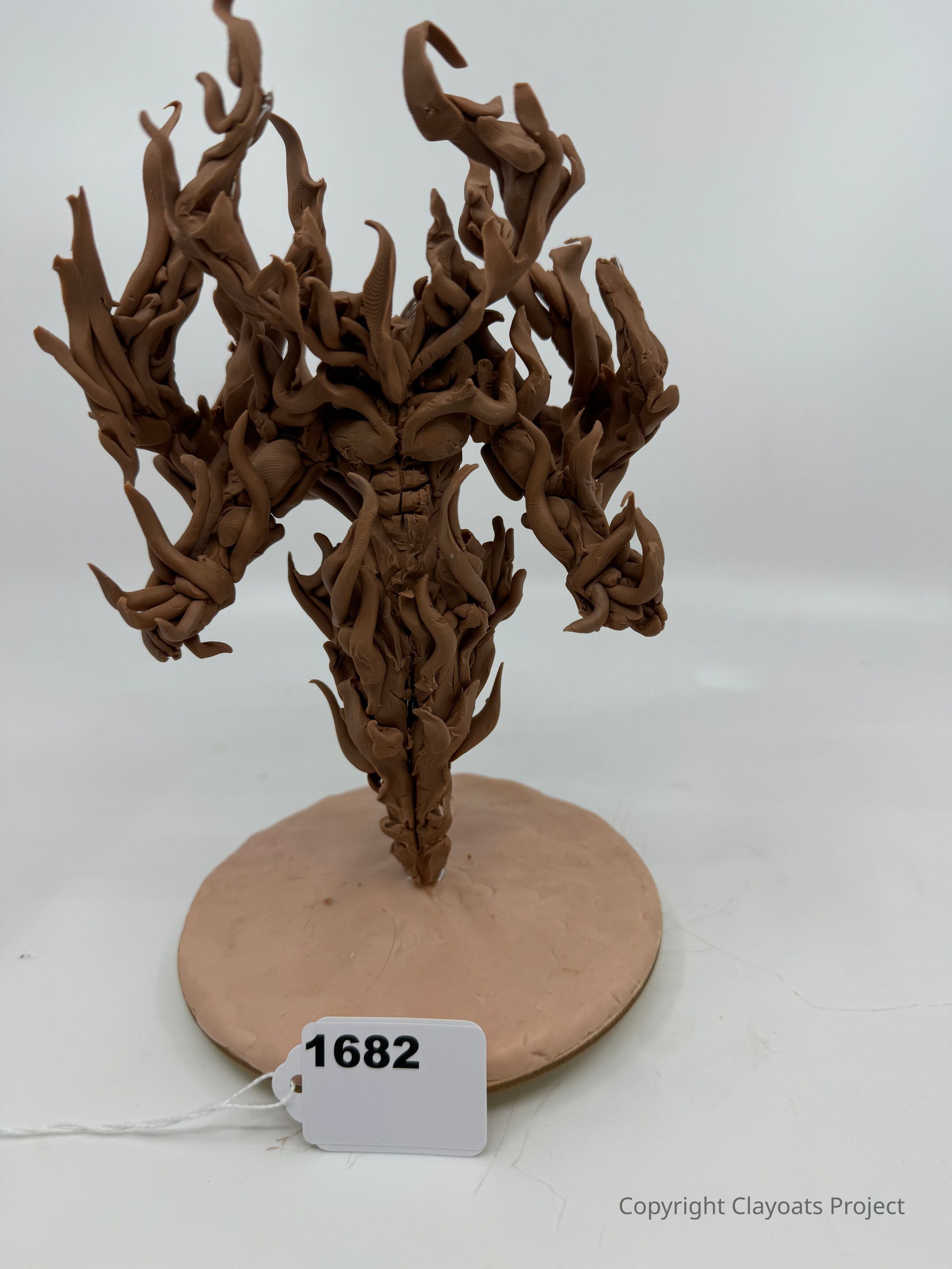 The image shows a clay sculpture of a humanoid figure with swirling, vine-like forms extending from its body, standing on a circular base. The clay is brown, and the figure appears dynamic and organic, as if composed of intertwining tendrils. The background is a plain light gray, which contrasts with the intricate details of the piece.