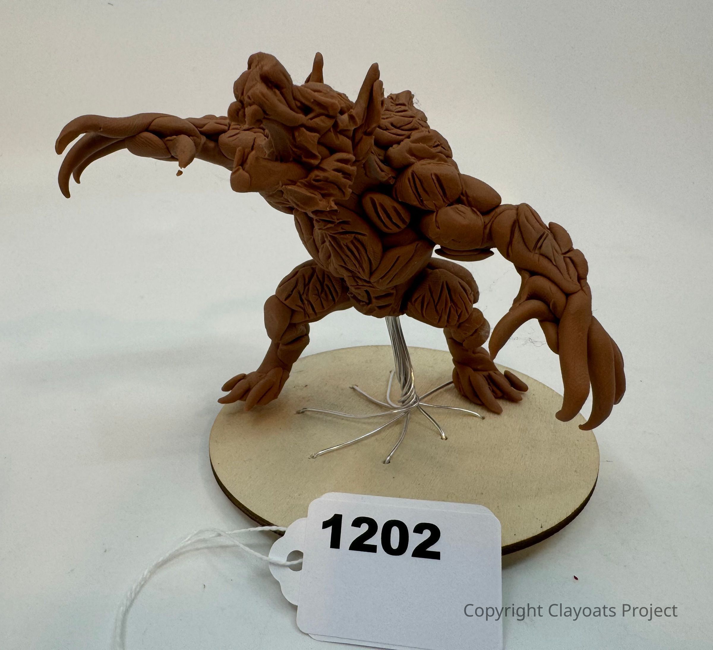 The image displays a detailed clay sculpture of a fantastical creature with expressive features, standing on a round wooden base, showcasing intricate textures and patterns on its body, limbs, and claws; which imbues the scene with an atmosphere of creativity and careful craftsmanship, as the wire support blends seamlessly into the design, allowing the sculpture to appear dynamic and poised, ready to spring into life