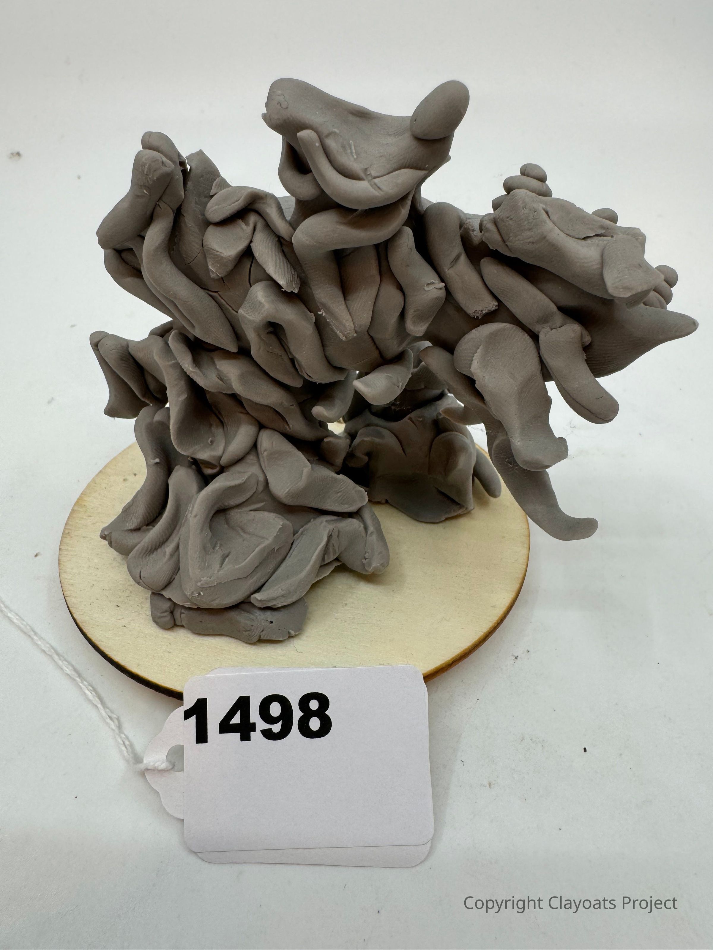 The image shows an abstract sculpture composed of twisted, gray clay forms with an organic appearance, mounted on a round wooden base. The sculpture appears dynamic and intricate, with various smooth, irregular shapes intertwined in a complex arrangement. The background is plain white, highlighting the structure and allowing focus on the sculpture’s details and texture without distractions.