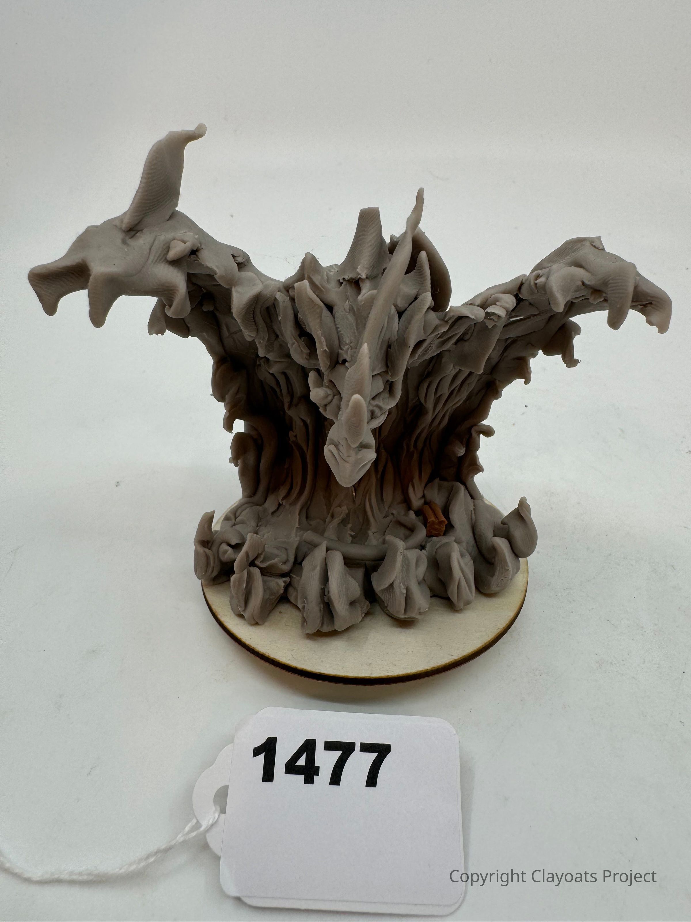 The image depicts a sculpture resembling a fantastical creature or abstract form with twisting, organic shapes and protrusions, rendered in a gray material and positioned on a circular base. Its surface has various textures, with some areas appearing smooth while others have more ridges and folds. The sculpture appears to capture movement, as if the form is dynamically emerging or unraveling. In front of the sculpture.