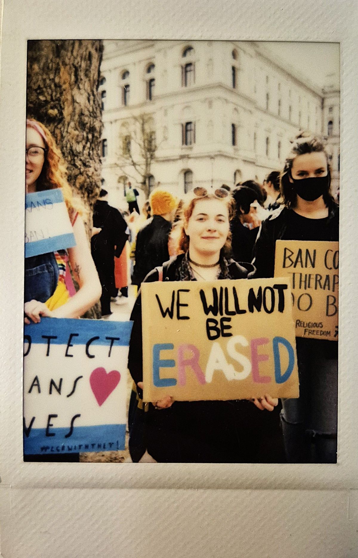A group of people hold signs at a protest, with supportive messages such as "WE WILL NOT BE ERASED".
