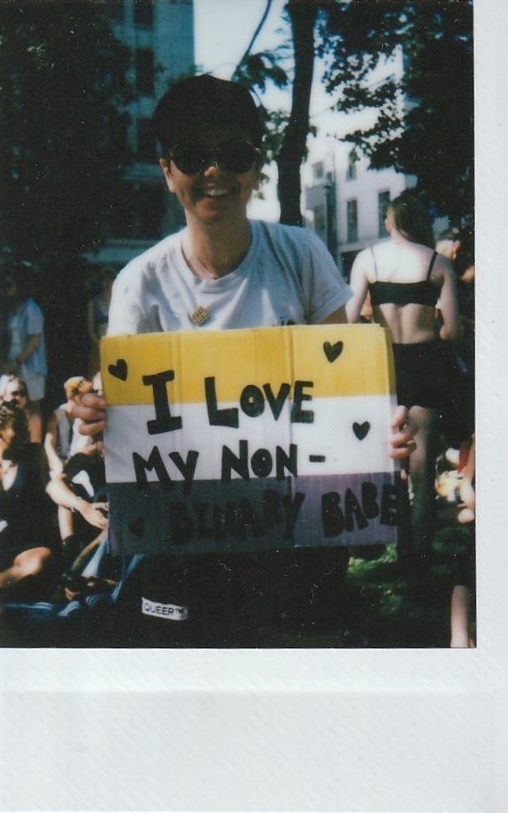 A person wearing sunglasses holds a sign reading, "I love my non-binary babe".