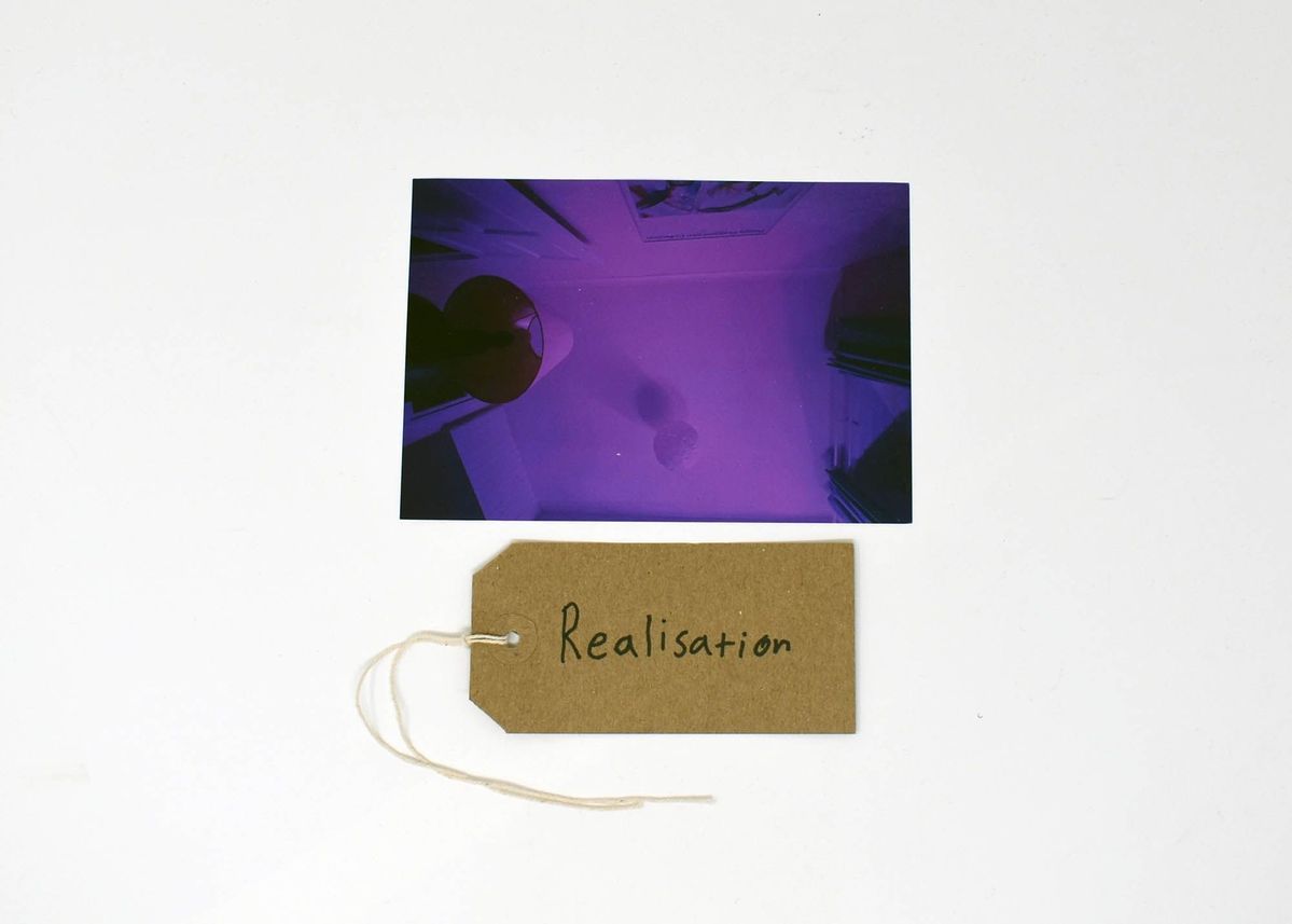 A purple photo of a ceiling with a brown handwritten luggage tag inscribed with "Realisation"