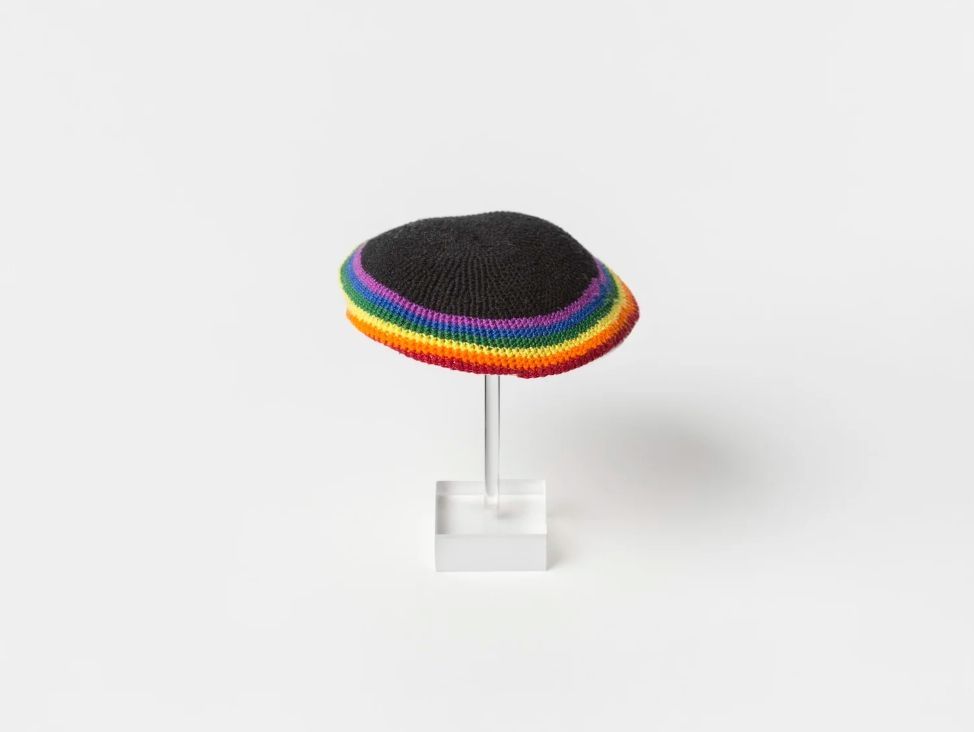 A rainbow knitted Kippah on a clear hat stand.