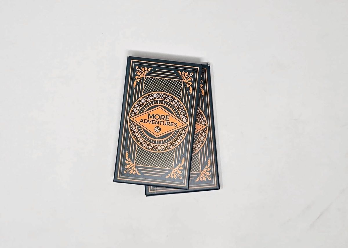 Small green box of cards with gold foil design reading "MORE ADVENTURES" inside a diamond.