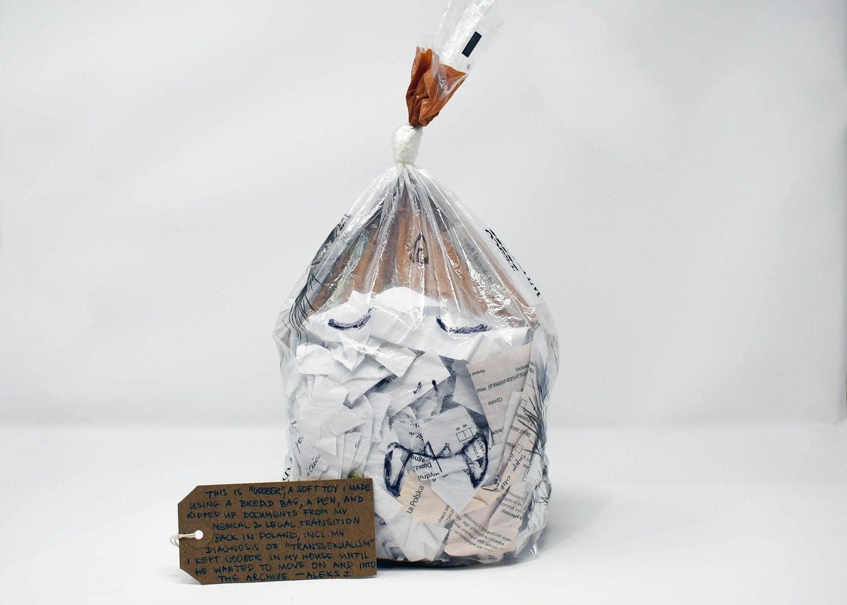 A clear plastic bread bag filled with ripped up paper documents and knotted. A cartoon face of a monster has been drawn on the bag. With a brown, handwritten luggage tag.