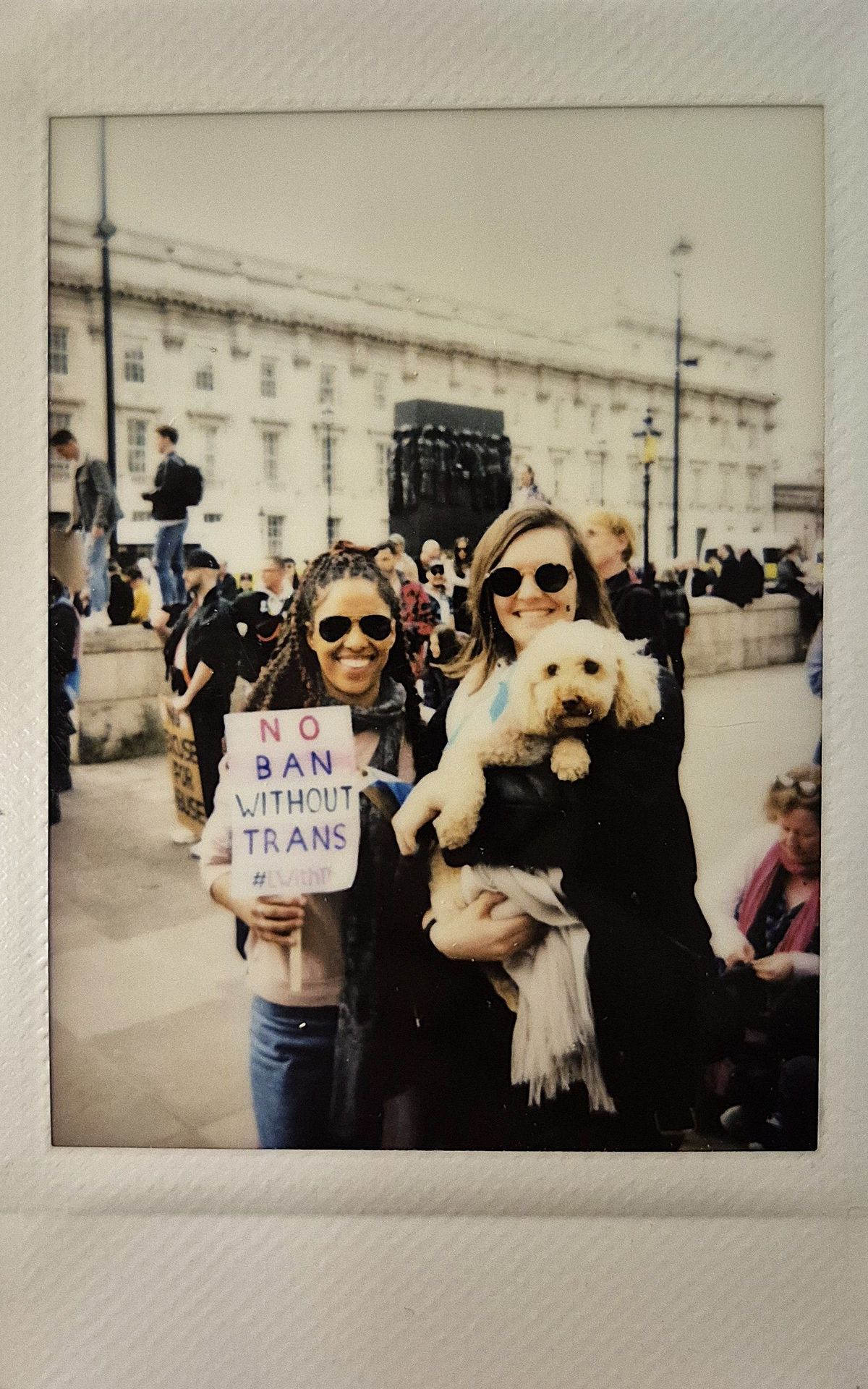 Two smiling individuals hold a small dog and a sign reading "No Ban Without Trans" during a public protest.