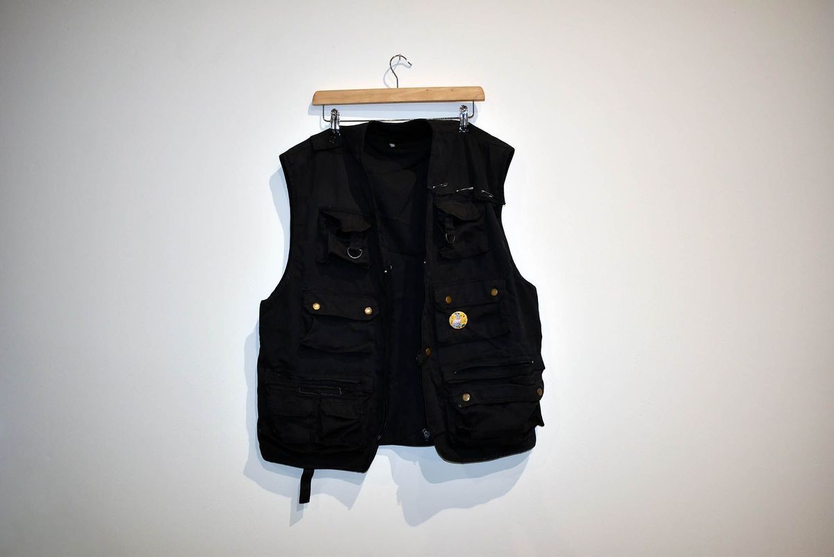 A black gilet with several pockets on a wooden clip hanger. It has a yellow badge (with a cat in the trans colours on it) pinned to the left breast pocket and is held together near the left shoulder with safety pins