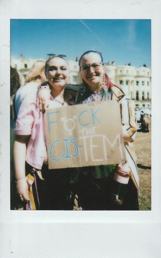 Two people at a protest are holding a cardboard sign which says "Fuck the Cis-Tem".