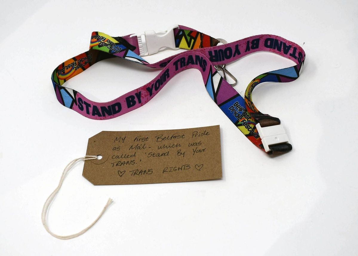 A colorful lanyard with “STAND BY YOUR TRANS” printed on it, with a brown luggage tag.