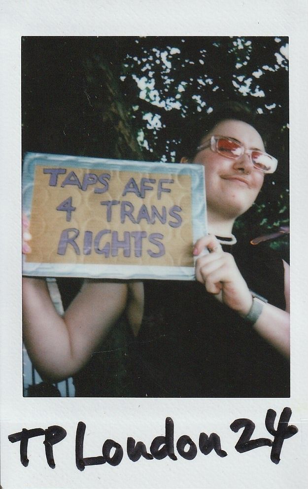 A person is holding a sign that reads "Taps Aff 4 Trans Rights" while smiling confidently