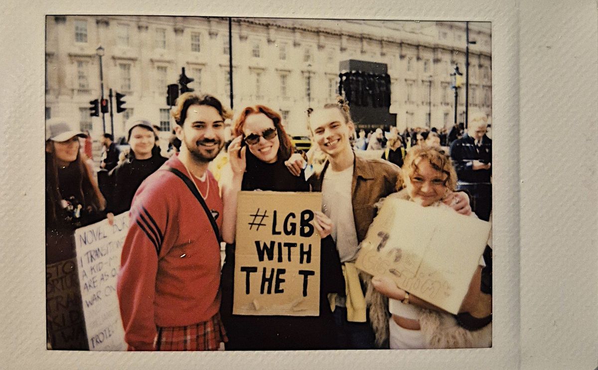 A group of people at a protest hold signs which say phrases such as "#LGB WITH THE T".