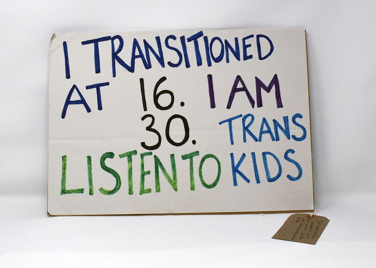 A white, cardboard placard with the text "I TRANSITIONED AT 16. I AM 30. LISTEN TO TRANS KIDS" in blue, black and green. With a brown, handwritten luggage tag.