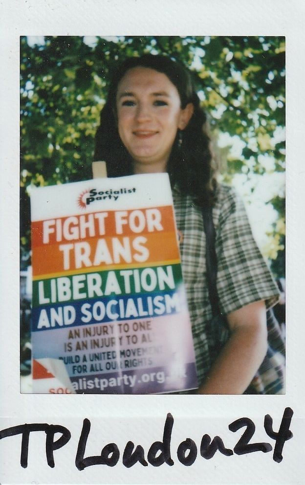 A person holds a colorful sign advocating for trans liberation and socialism at an outdoor event beneath trees.