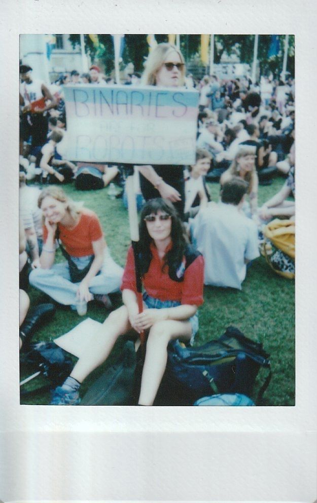 A group of people sits on grass at a protest holding a sign that reads "Binaries Are For Robots".