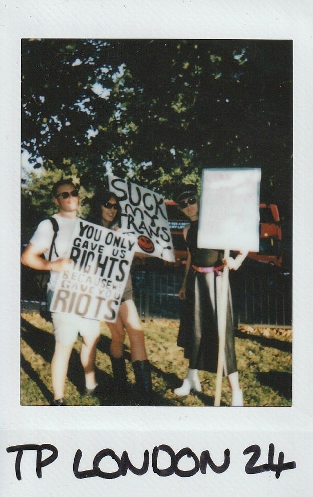 Three people stand holding protest signs in a sunlit park setting, holding placards.