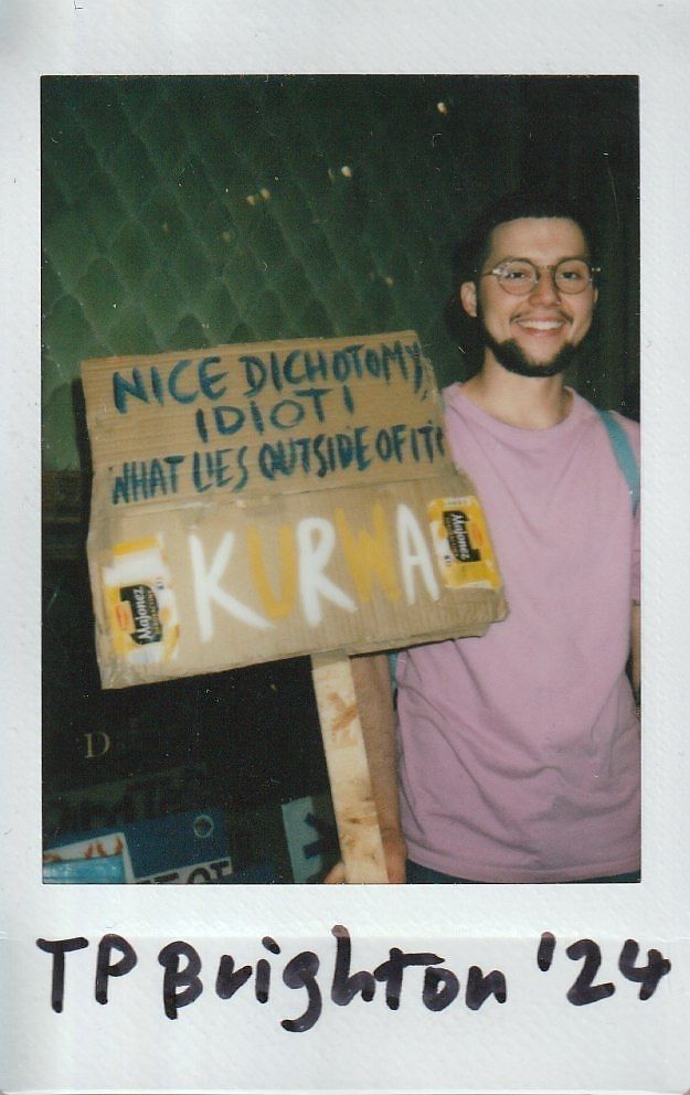 A smiling person holds a cardboard sign with various messages, standing against a textured background in TP Brighton.