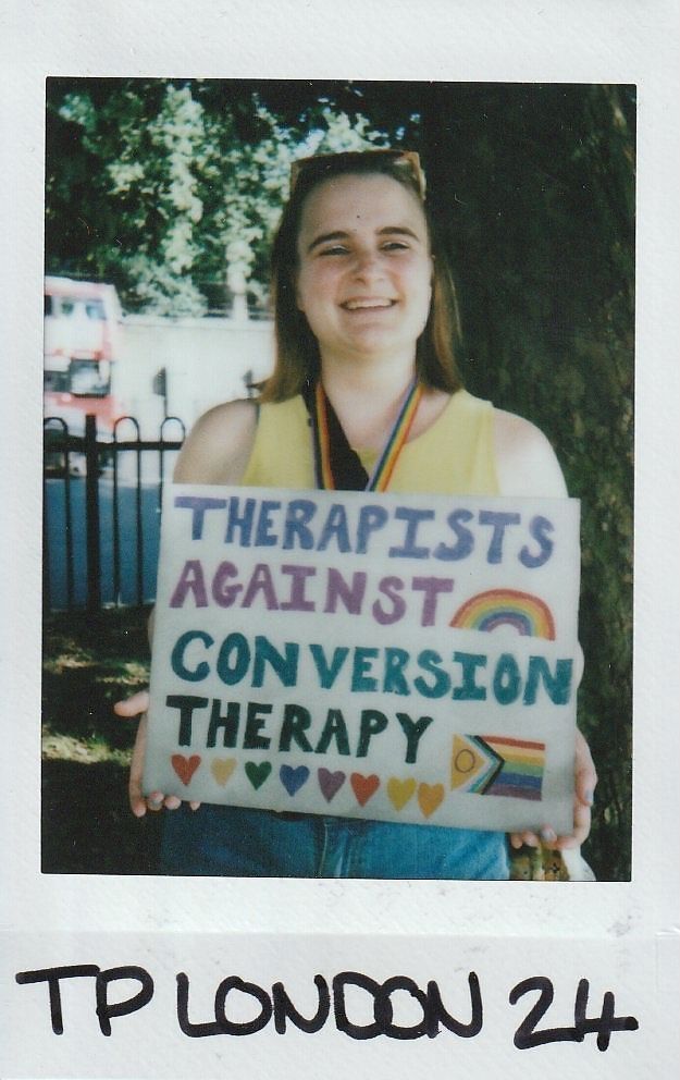 A person smiles while holding a sign stating "Therapists Against Conversion Therapy" with colorful decorations.