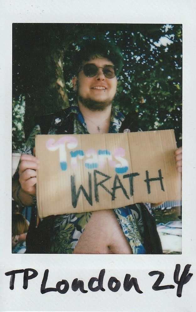 A person smiles while holding a sign saying "Trans WRATH" in a sunny outdoor setting.