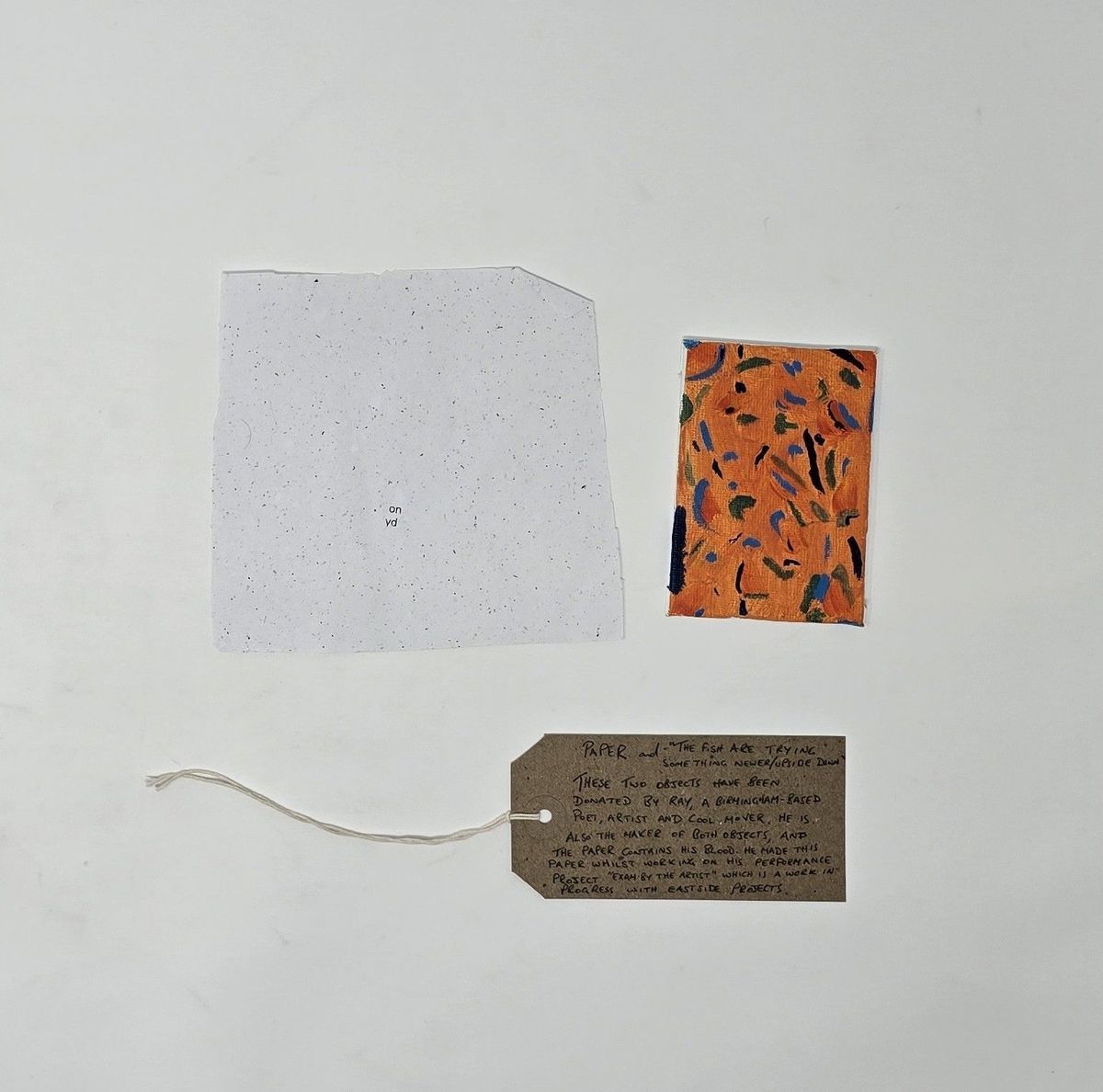 A white handmade piece of paper, a small red painting and a brown tag with writing on it lay on a white surface.