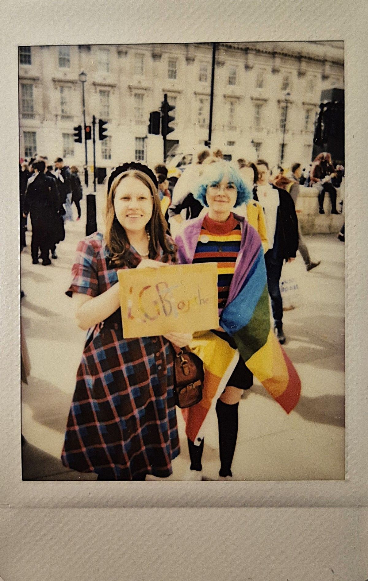 Two people stand smiling, one has a rainbow flag wrapped around them and the other is holding a small placard which says 'LGBTOGETHER'.