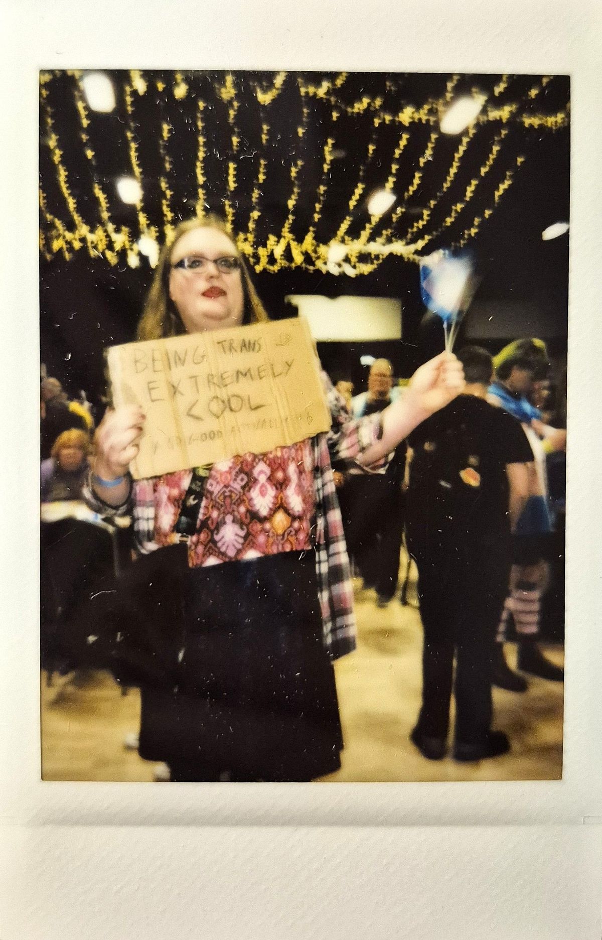 A person holds a sign reading "Being trans is extremely cool" in a lively, festive event space.