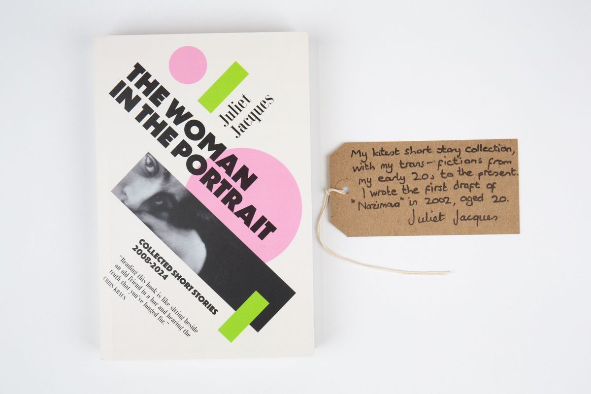 A book titled "The Woman in the Portrait - Collected Short Stories 2008-2024" by Juliet Jacques. The cover is white with green and pink geometric shapes and black and white close up photo of a person's eyes. With a brown, handwritten luggage tag.
