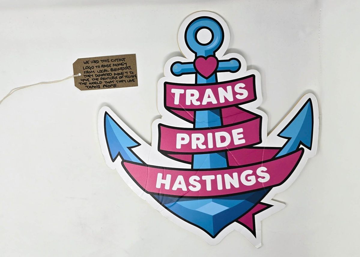 Image of a foam board cut out of the Trans Pride Hastings logo. The logo is a blue anchor with a pink heart a the top and the words "Trans Pride Hastings" in white on a pink ribbon entwined round the anchor. With a brown, handwritten luggage tag.