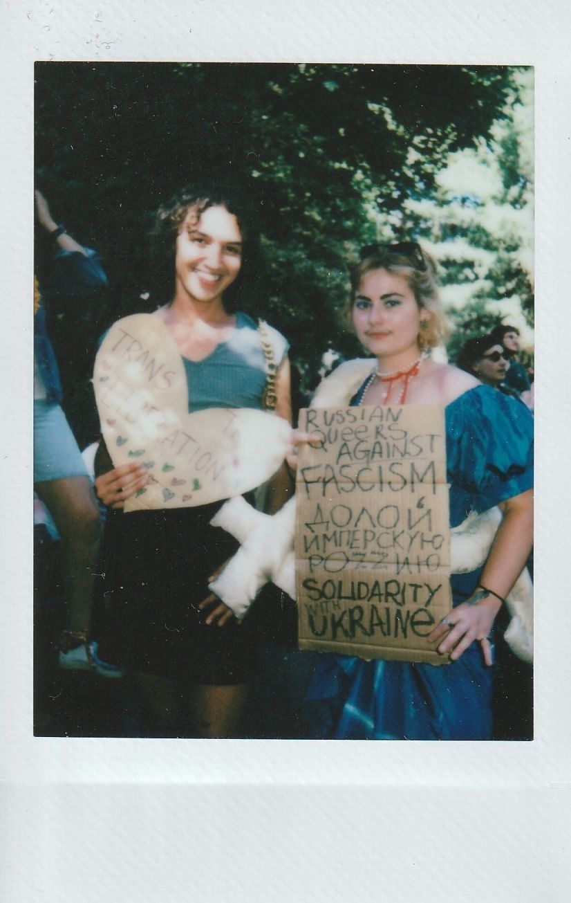 Two people holding signs, one which says "Trans Liberation" and the other "Russian queers against fascism solidarity with Ukraine".