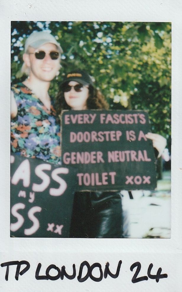Two people holding a handmade sign with a message stand together outdoors, surrounded by greenery.