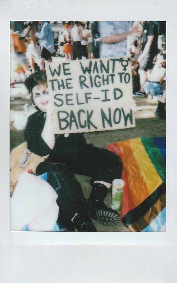 A person sits on the ground holding a sign which says "WE WANT THE RIGHT TO SELF-ID BACK NOW"