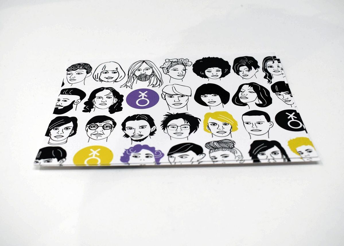 Triangular scarf laying on a wooden surface with point at the bottom. Across the top of the scarf is a black stripe with white block text reading "IF WE ARE NOT VISIBLE WE CANNOT DEMAND THE WORLD MAKE SPACE." The rest of the scarf shows 4 rows of simple line-art drawings of various faces, occassionally interspersed with the non-binary symnol on purple and yellow circles.