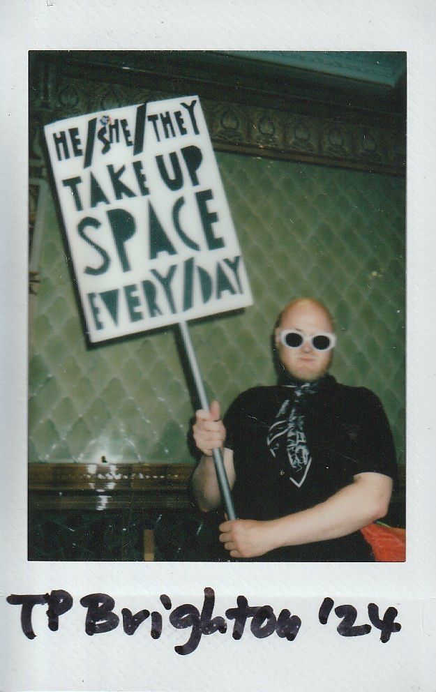 A person holding a sign advocating inclusivity stands confidently in a room, wearing sunglasses and a patterned scarf.
