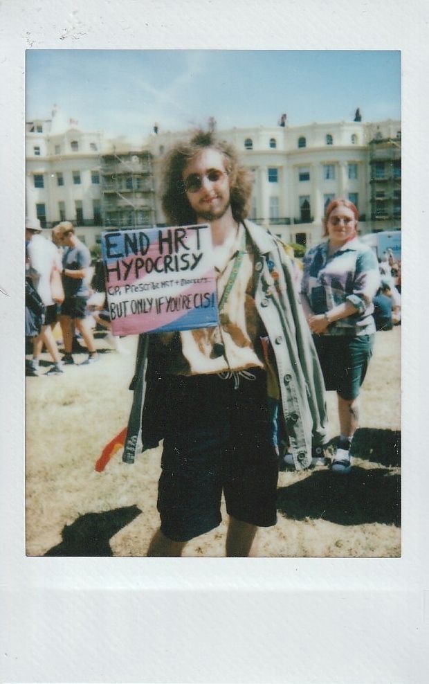 A person stands on grass, holding a sign about "END HRT hypocrisy".