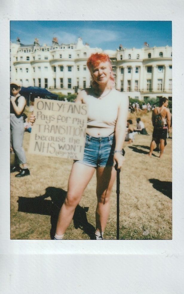 A person stands outside with a cane, holding a sign which says "Only Fans Pays For My Transition Because The NHS Won't".