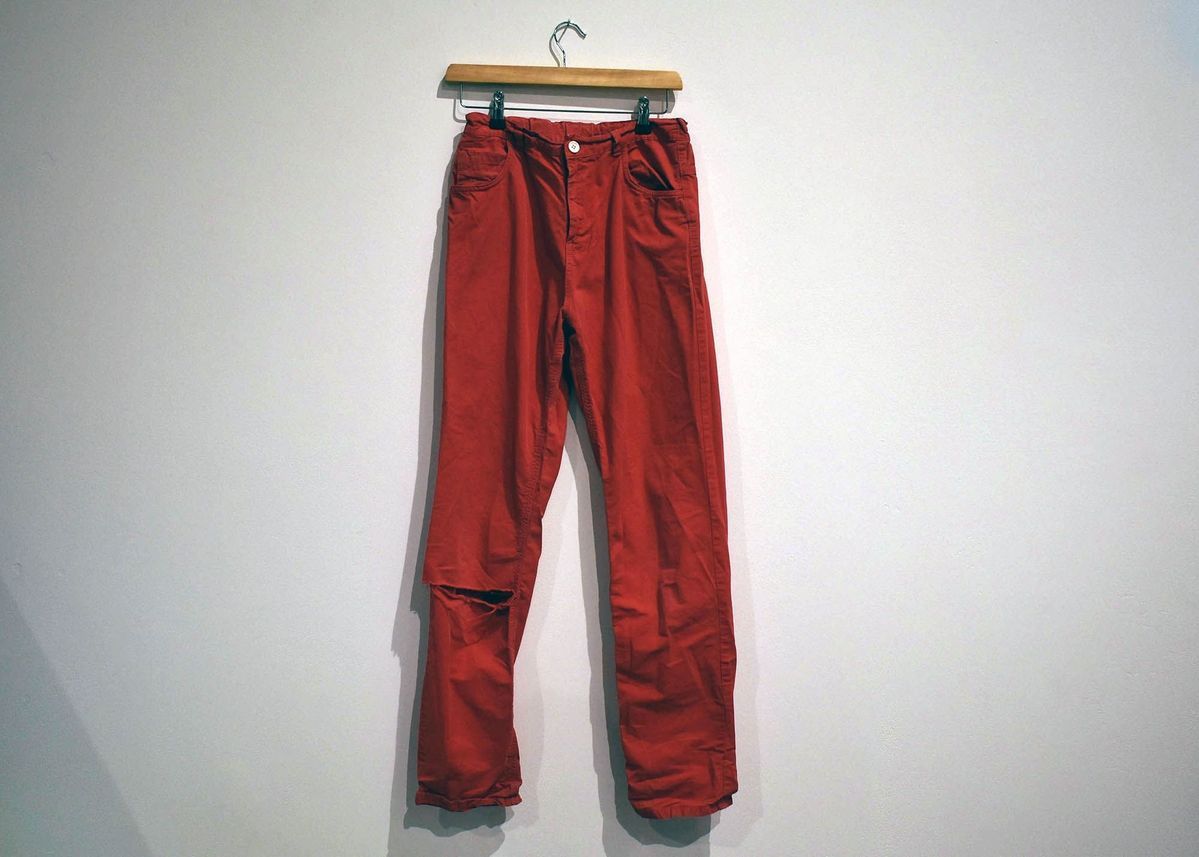 A pair of red pants is hanging on a wooden hanger against a plain wall.