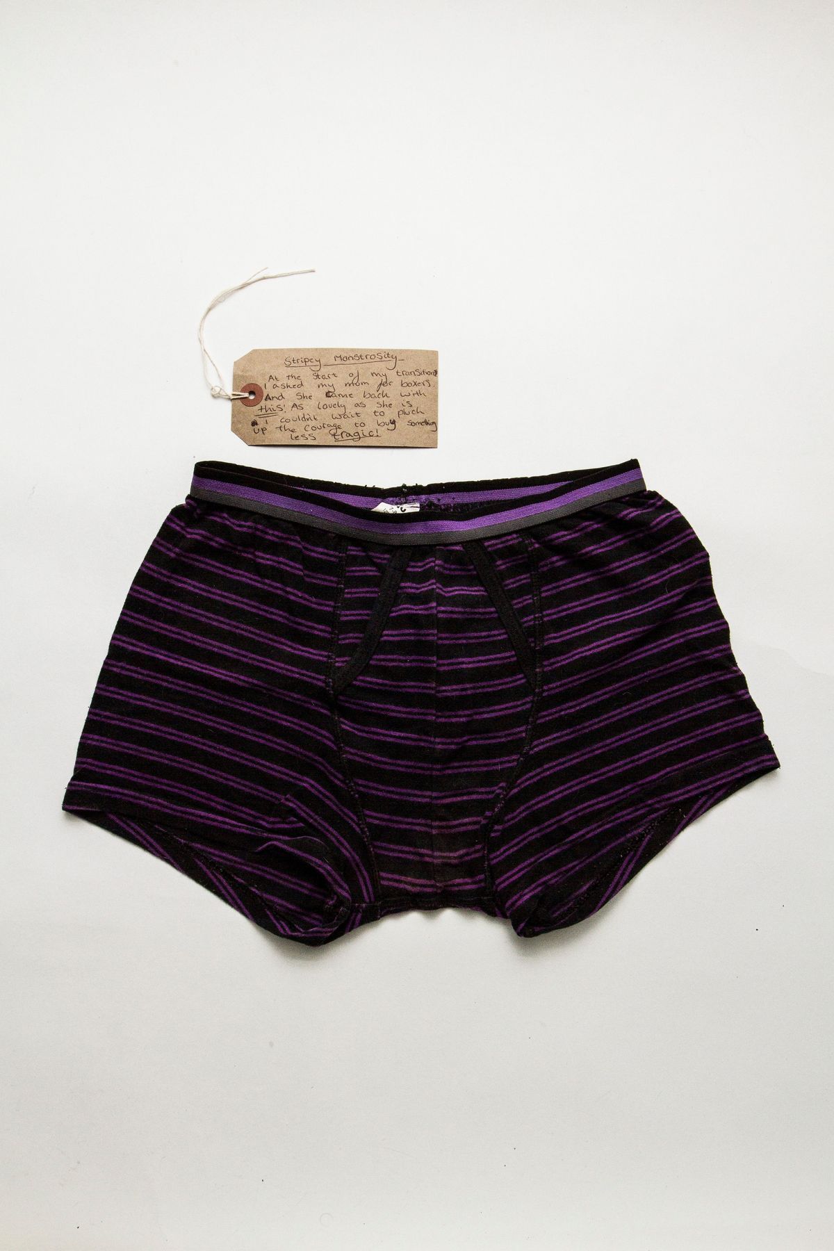 A pair of boxer shorts with purple and black narrow stripes. The boxer shorts are in worn condition and some fraying can be seen in places.