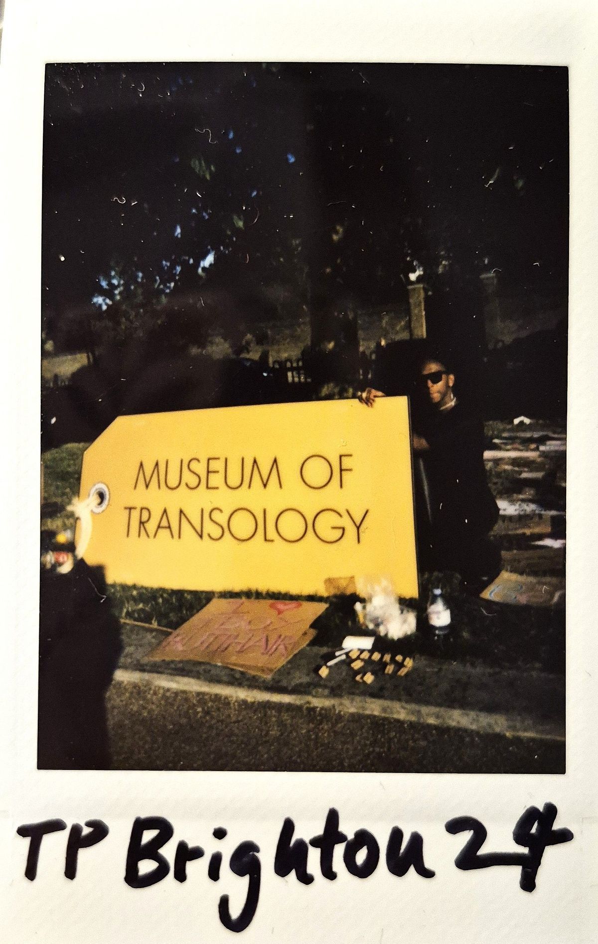 A person poses beside a large, yellow sign labeled "Museum of Transology," with various items nearby.