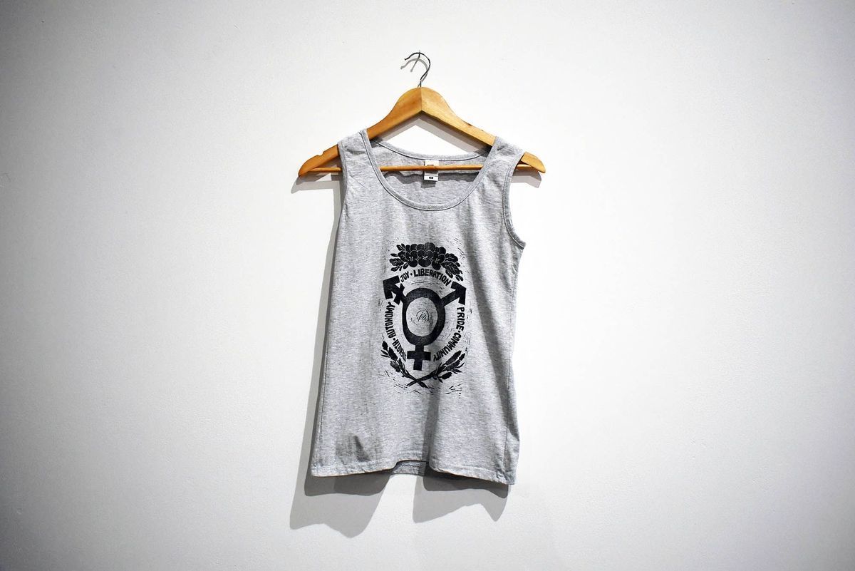 A grey tank top printed in black with the trans symbol surrounded by the words STRENGTH AUTONOMY JOY LIBERATION PRIDE COMMUNITY and floral motifs, on a wooden hanger.