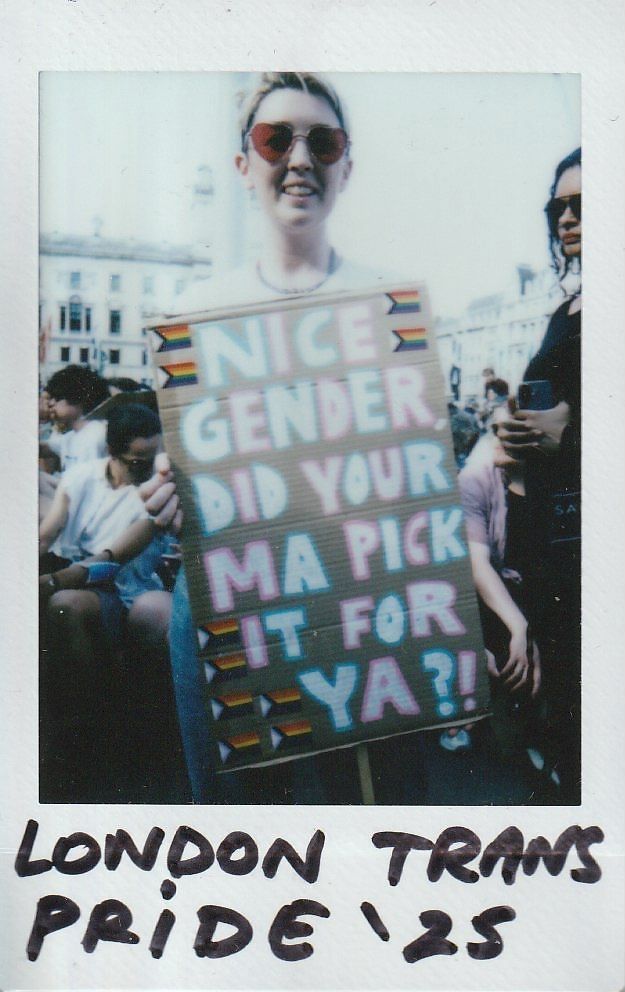 A person smiles, holding a sign at London Trans Pride which says "Nice Gender Did Your Ma Pick It For Ya?!"