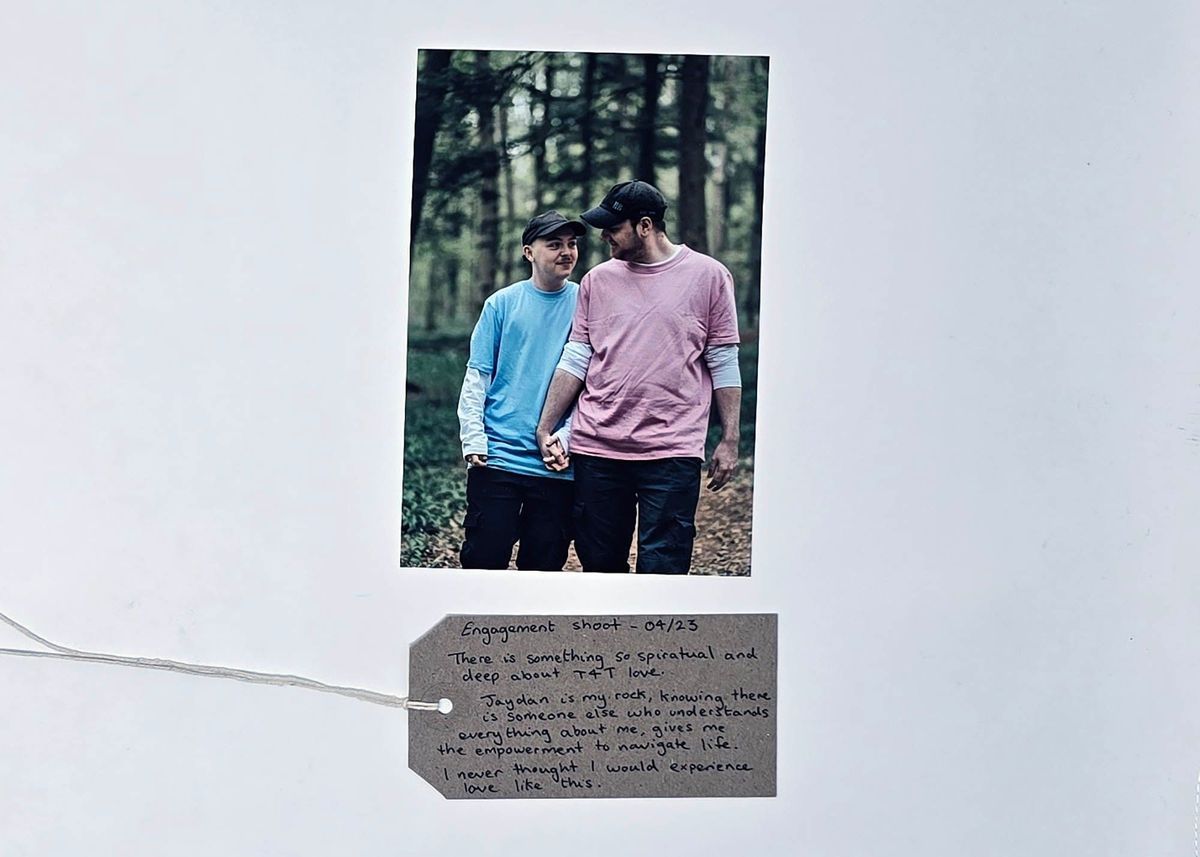A photo of two people holding hands, and each others gaze, in a wood celebrating their engagement. Both wear baseball caps, one a blue t-shirt, the other a pink t-shirt. With a brown handwritten luggage tag.