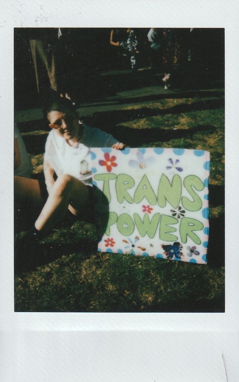 A person is sitting on grass, holding a colorful sign that says “Trans Power” in large letters.