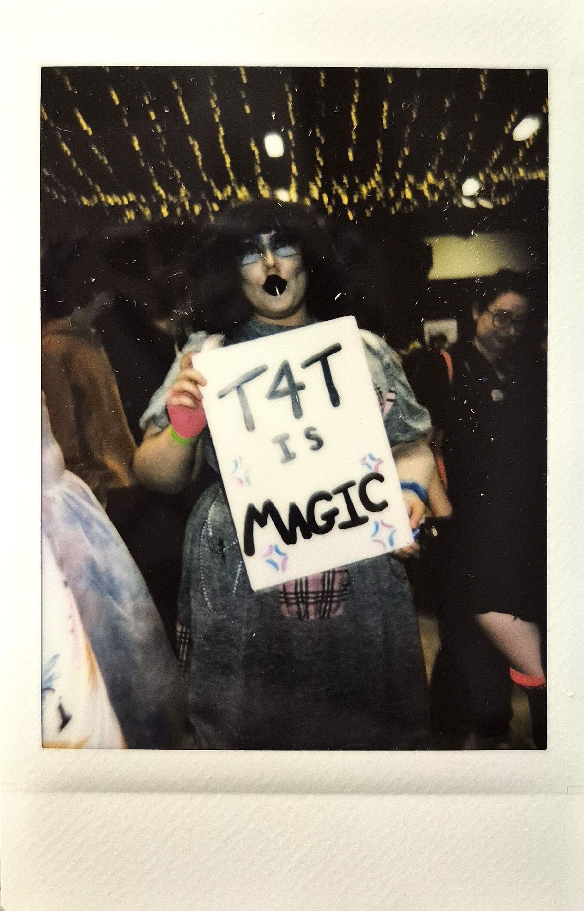 A person holds a sign saying “T4T is magic” with colorful decorations underneath vibrant lights.