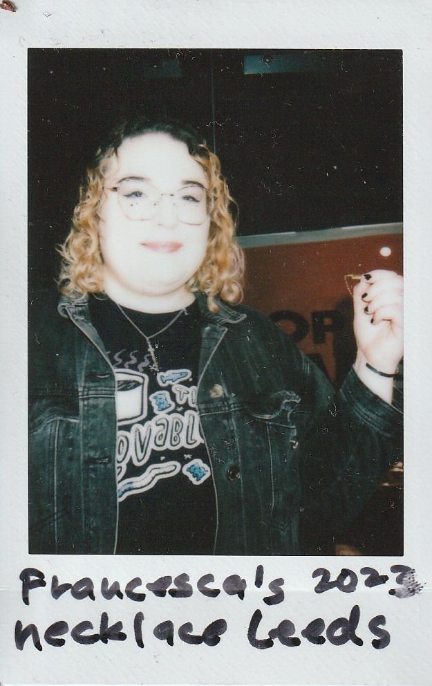 A person with curly hair is smiling, wearing glasses, and holding a necklace card in indoor setting.