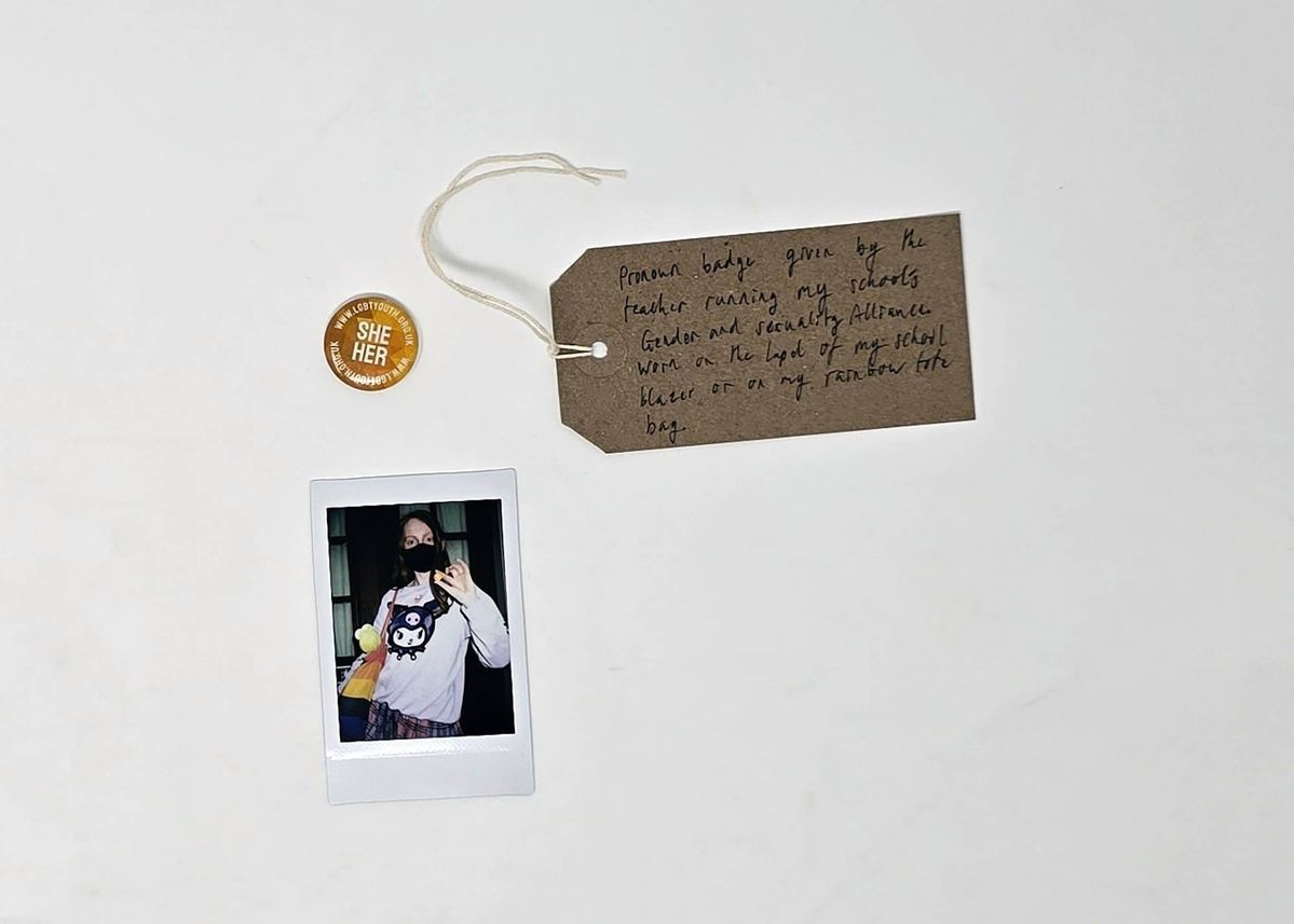 A small, orange, "SHE HER" pronoun pin badge from LGBTYOUTH. With a Polaroid and a brown, handwritten luggage tag.