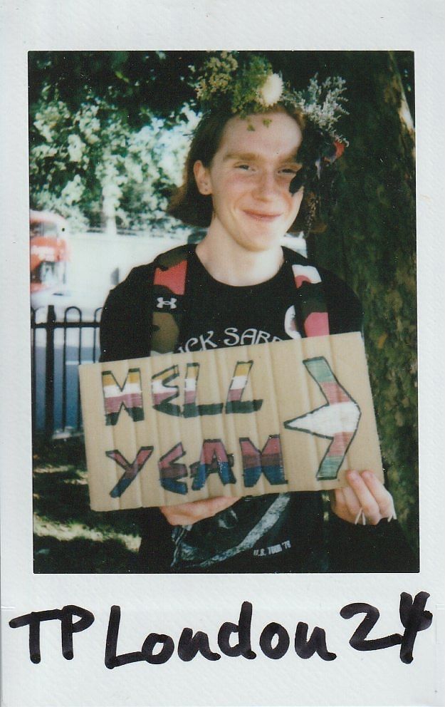 A person smiles while holding a colorful sign saying "HELL YEAH," wearing a floral crown.
